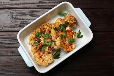 Photo of Tasty cauliflower steaks and parsley in baking dish on wooden table, top view