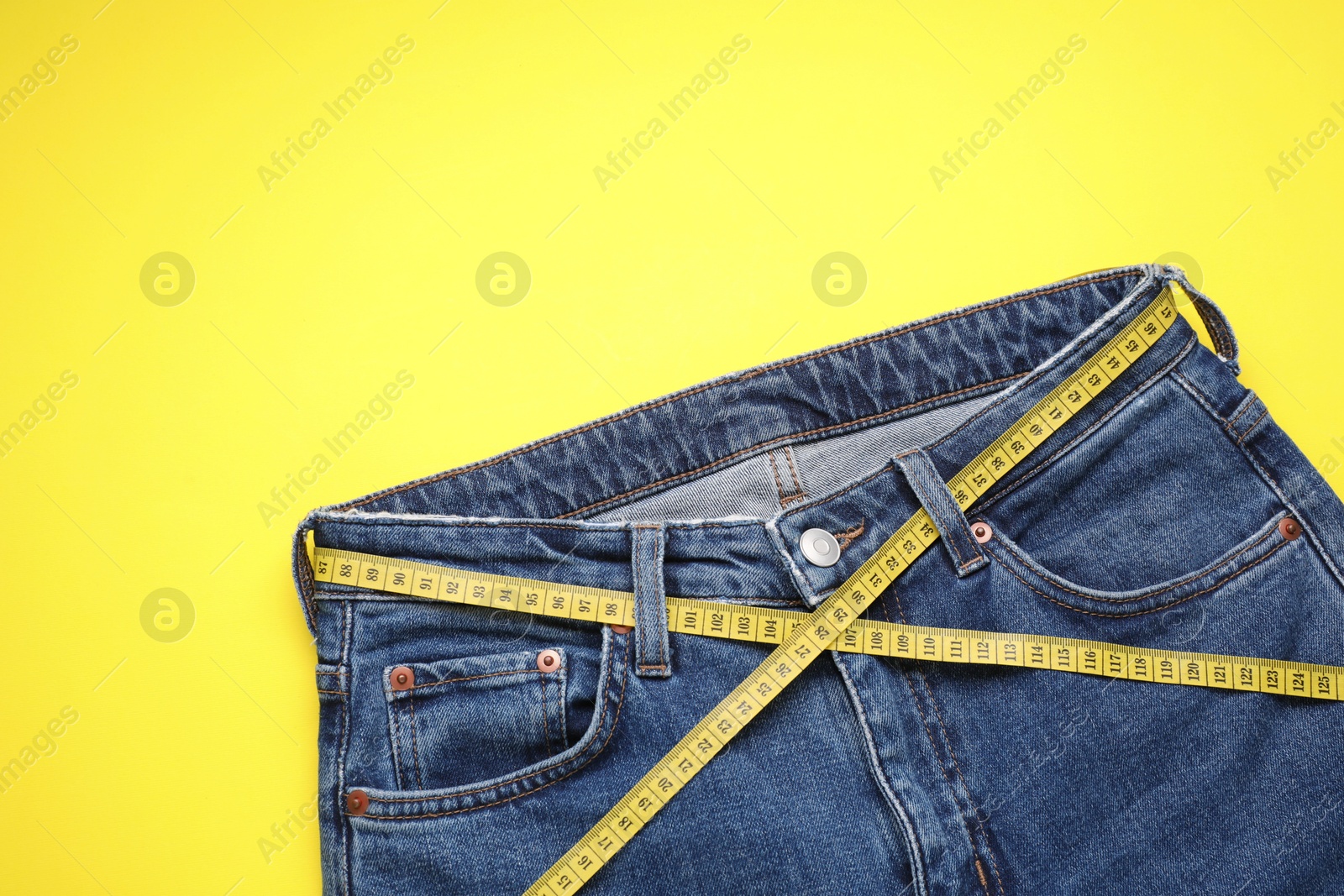 Photo of Jeans and measuring tape on yellow background, top view. Space for text