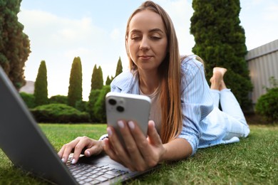 Woman with smartphone using laptop on green lawn in park