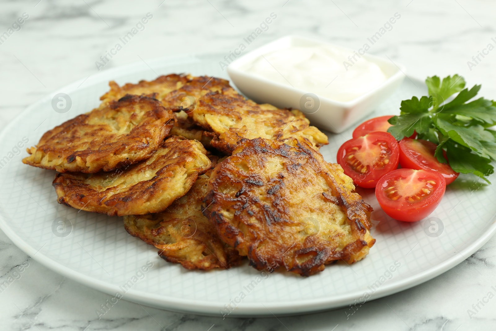 Photo of Delicious potato pancakes with fresh tomatoes, sour cream and parsley on white marble table, closeup