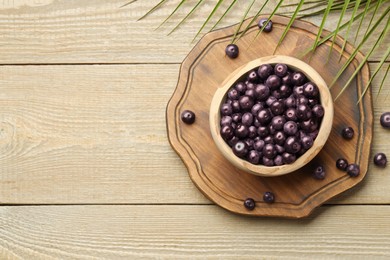 Photo of Ripe acai berries in bowl and palm leaves on wooden table, flat lay. Space for text