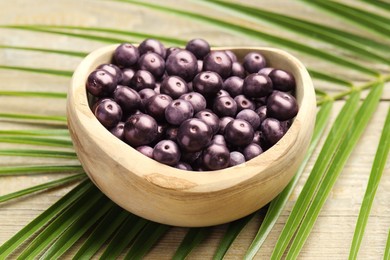 Photo of Ripe acai berries in bowl and palm leaves on wooden table, closeup