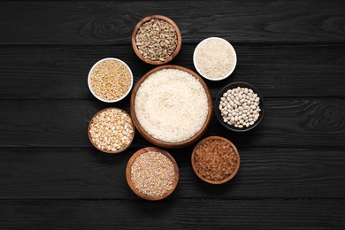 Photo of Different types of cereals, seeds and legumes in bowls on black wooden table, flat lay