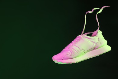 Photo of Stylish sneaker in air against dark green background in neon lights, space for text