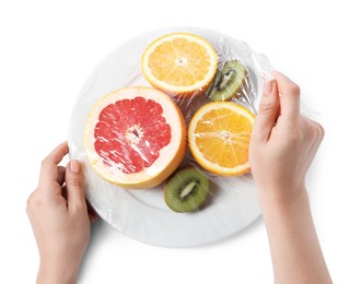 Woman putting plastic food wrap over plate with fresh fruits on white background, top view