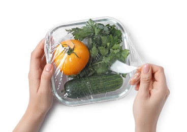 Woman putting plastic food wrap over glass container with vegetables on white background, top view