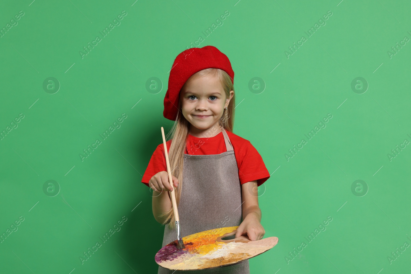 Photo of Little girl with brush and palette pretending to be artist on green background. Dreaming of future profession