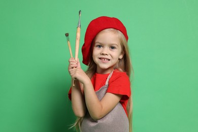 Little girl with brushes pretending to be artist on green background. Dreaming of future profession