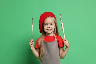 Little girl with brushes pretending to be artist on green background. Dreaming of future profession