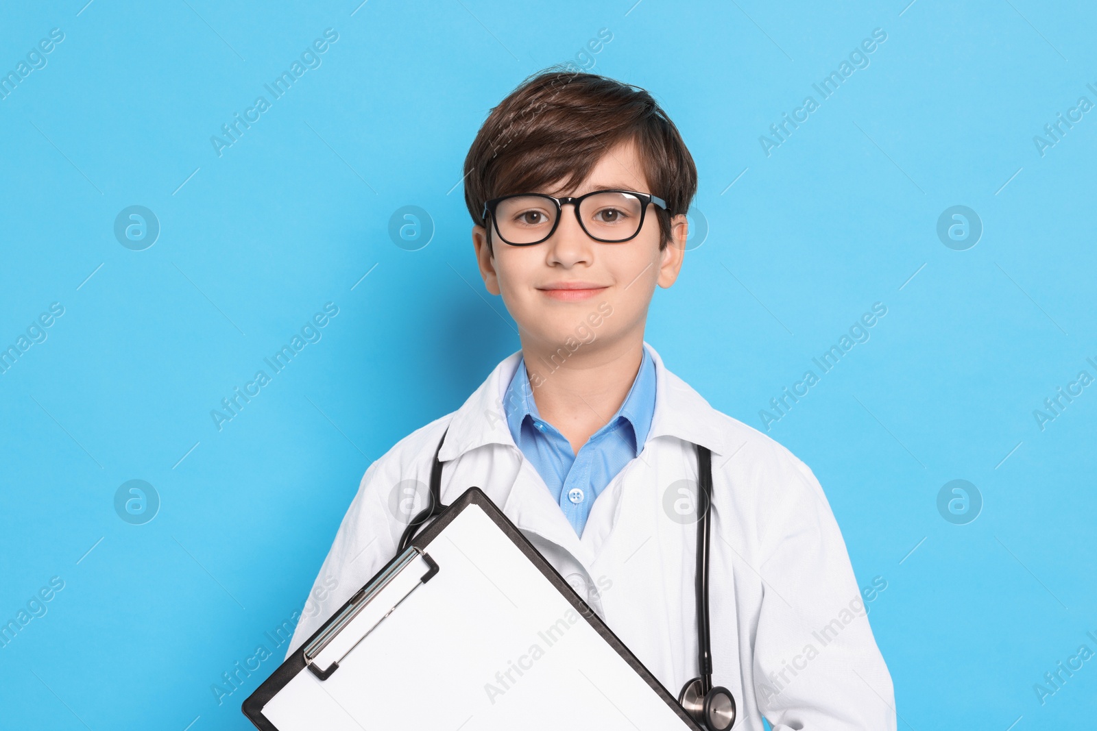 Photo of Boy with stethoscope and clipboard pretending to be doctor on light blue background. Dreaming of future profession