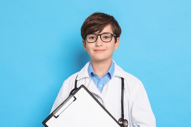 Photo of Boy with stethoscope and clipboard pretending to be doctor on light blue background. Dreaming of future profession