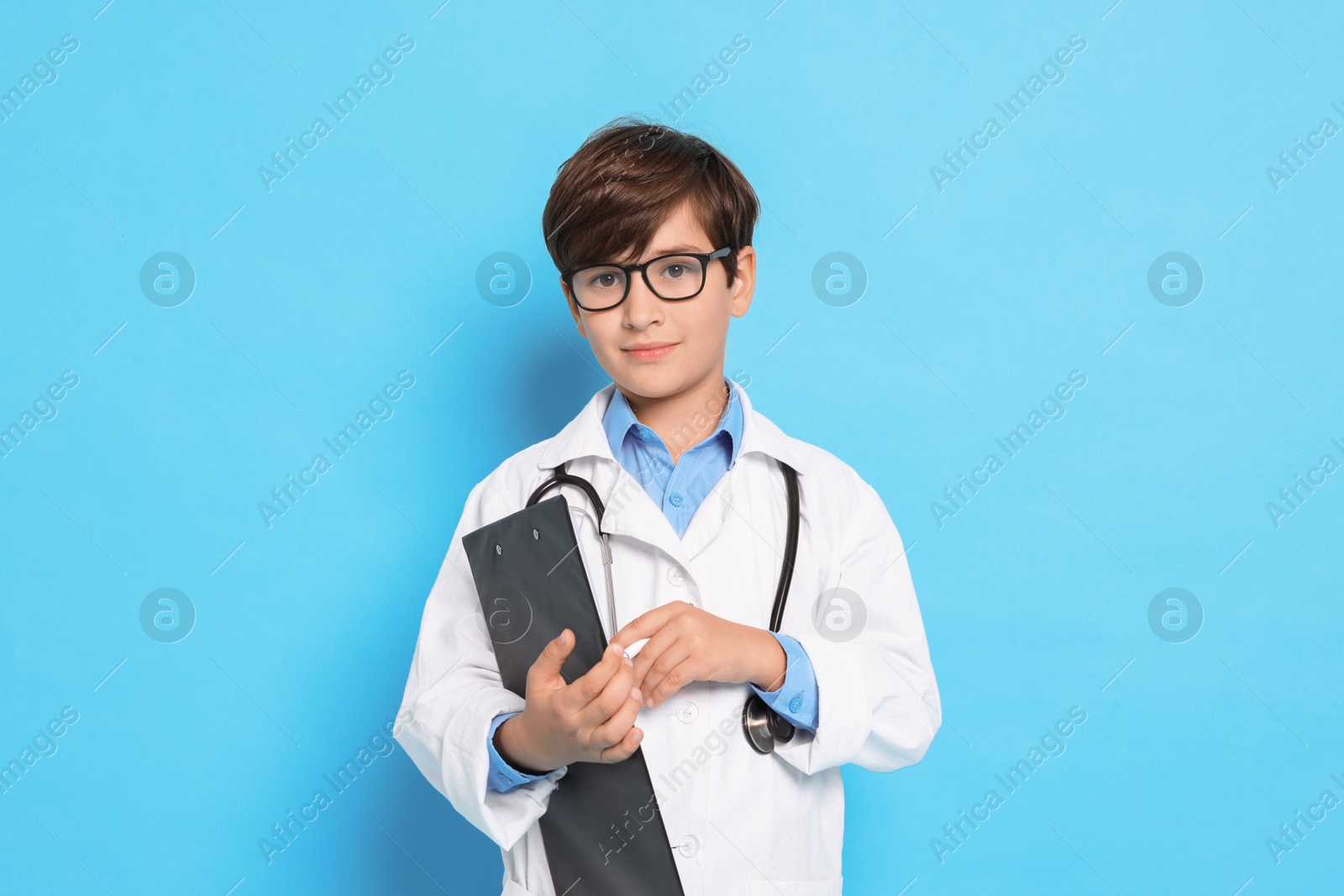 Photo of Boy with stethoscope and clipboard pretending to be doctor on light blue background. Dreaming of future profession