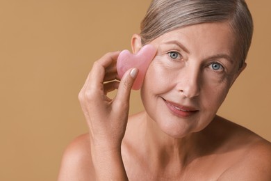 Beautiful woman doing facial massage with gua sha tool on beige background, closeup