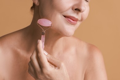 Woman doing facial massage with roller on beige background, closeup