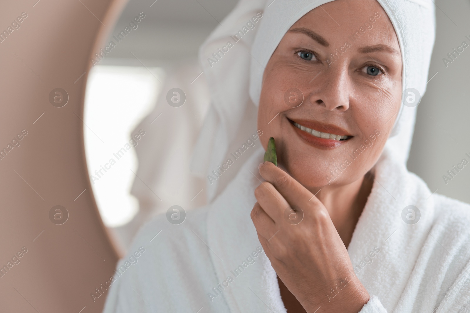 Photo of Beautiful young woman doing facial massage with gua sha tool indoors