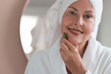 Beautiful young woman doing facial massage with gua sha tool indoors