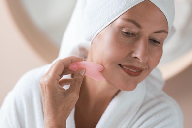 Photo of Beautiful young woman doing facial massage with gua sha tool indoors