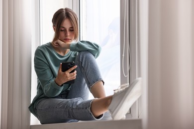 Photo of Loneliness concept. Sad teenage girl using smartphone on windowsill at home, low angle view