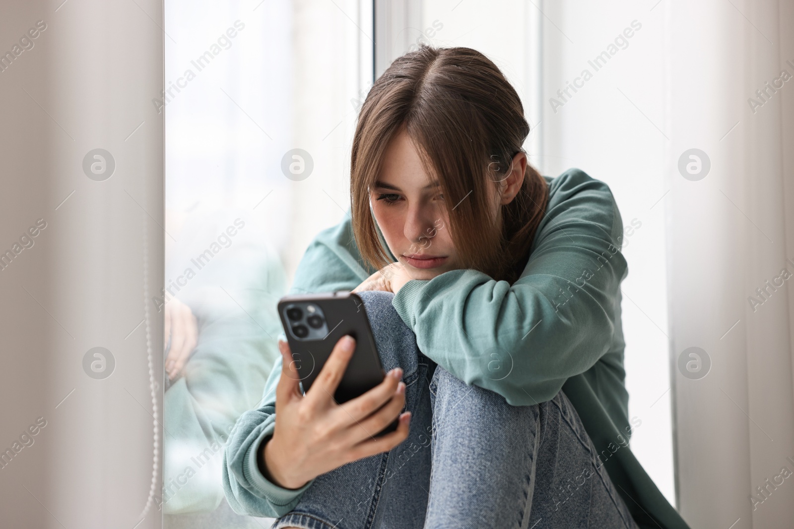 Photo of Loneliness concept. Sad teenage girl using smartphone on windowsill at home