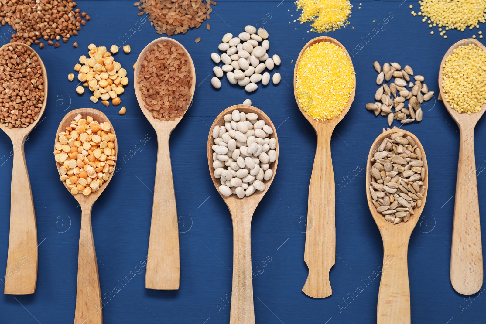 Photo of Different types of cereals, seeds and legumes in spoons on blue wooden table, flat lay