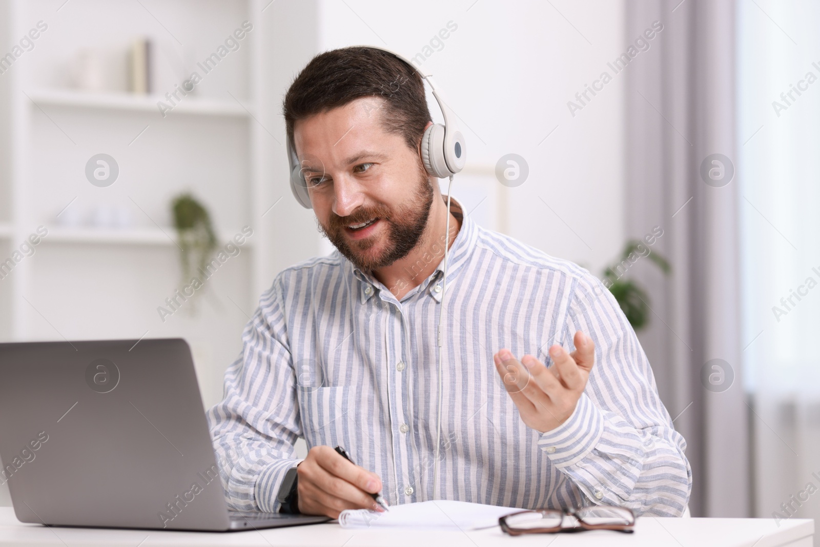 Photo of Interpreter in headphones taking notes while having video chat via laptop at white table indoors