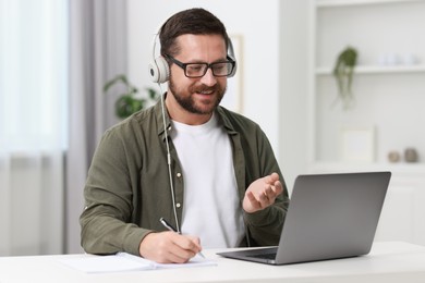 Interpreter in headphones taking notes while having video chat via laptop at white table indoors