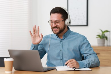 Photo of Interpreter in headphones taking notes while having video chat via laptop at wooden table indoors