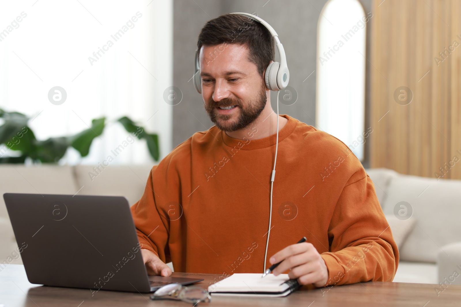 Photo of Interpreter in headphones taking notes while having video chat via laptop at wooden table indoors