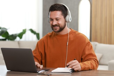 Photo of Interpreter in headphones taking notes while having video chat via laptop at wooden table indoors