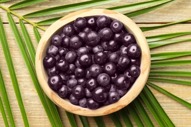 Photo of Ripe acai berries in bowl and palm leaves on wooden table, top view