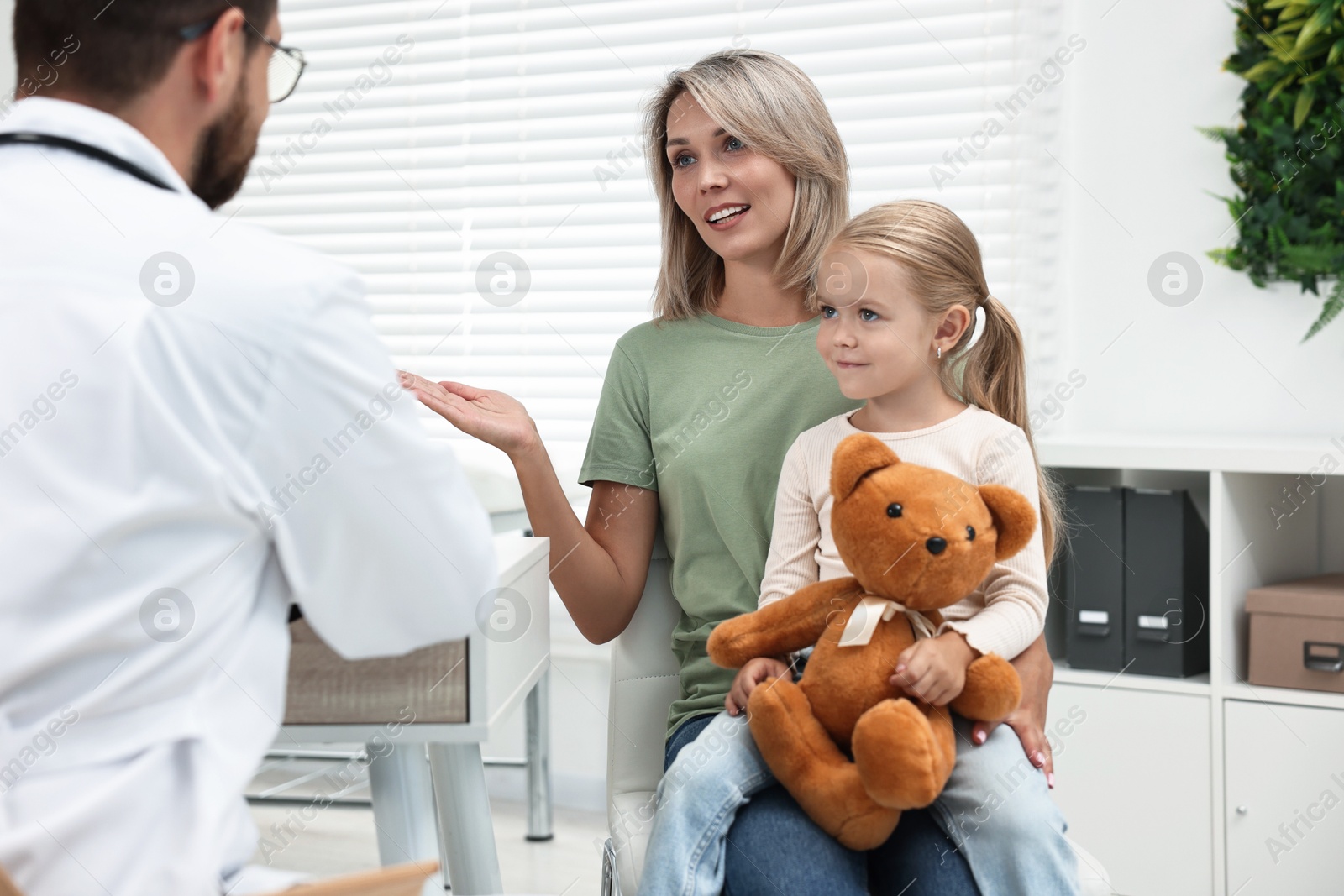 Photo of Doctor consulting little girl with teddy bear and her mother in hospital, back view