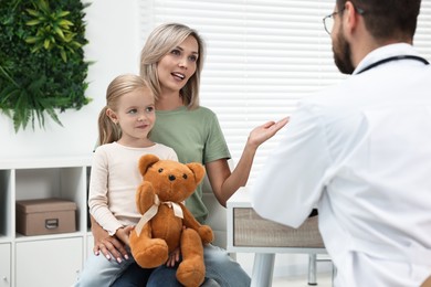 Photo of Doctor consulting little girl with teddy bear and her mother in hospital, back view