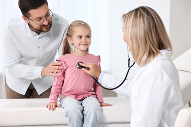 Photo of Doctor examining little girl with stethoscope and her father in hospital