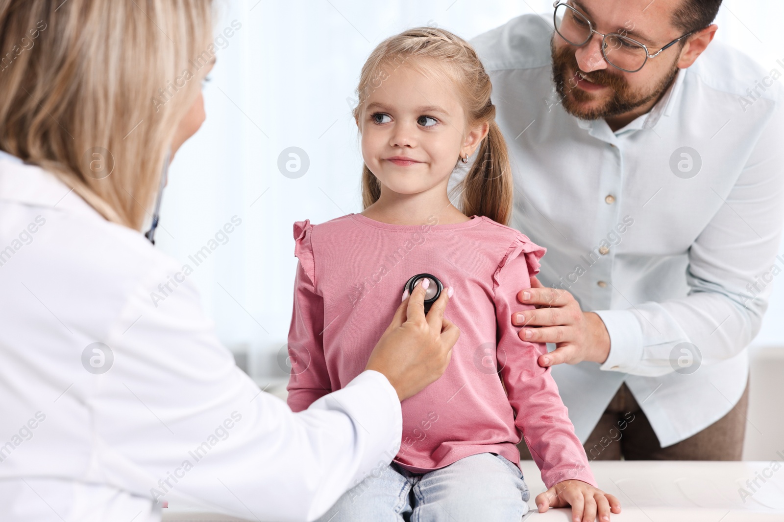 Photo of Doctor examining little girl with stethoscope and her father in hospital