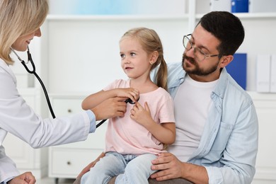 Photo of Doctor examining little girl with stethoscope and her father in hospital