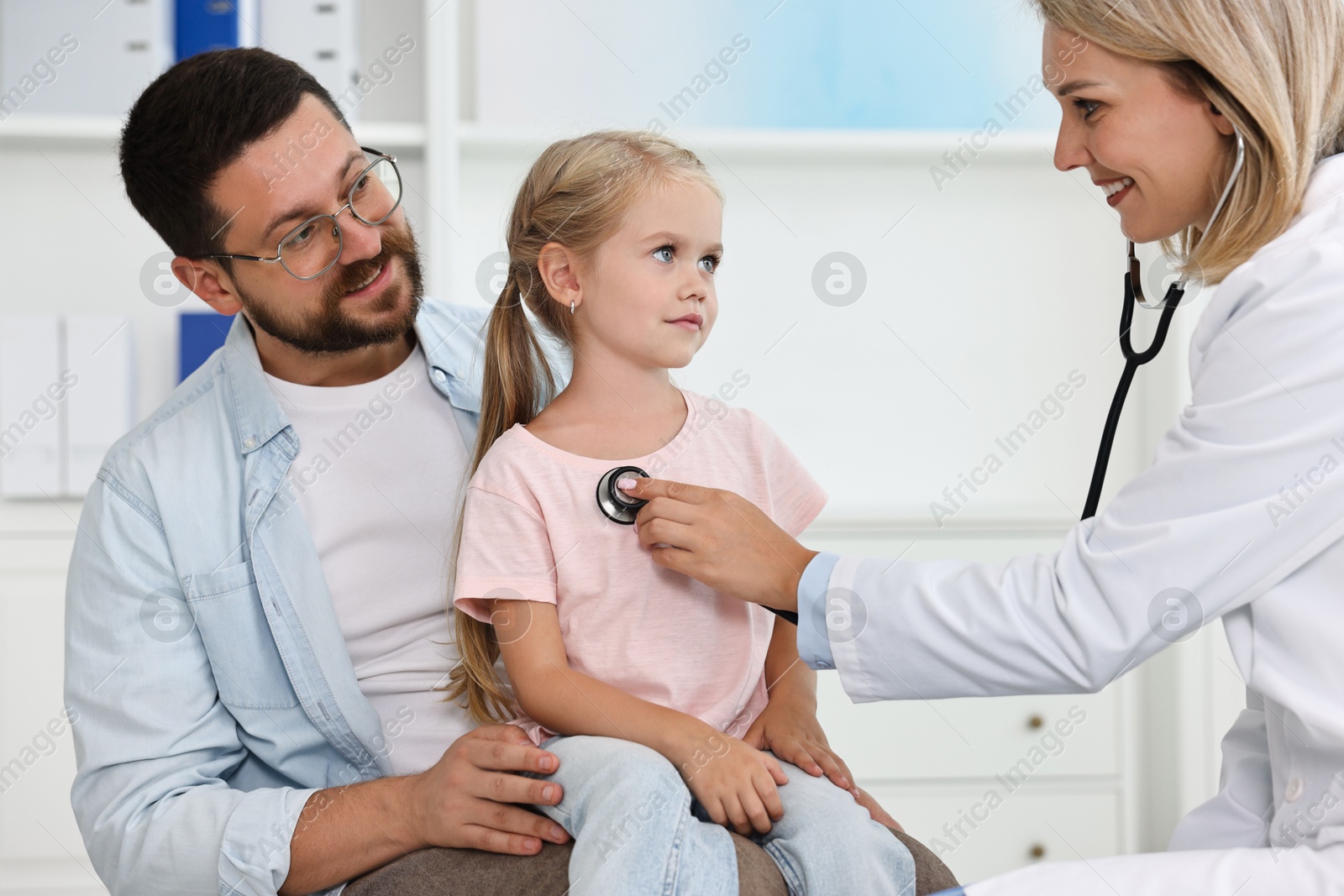 Photo of Doctor examining little girl with stethoscope and her father in hospital