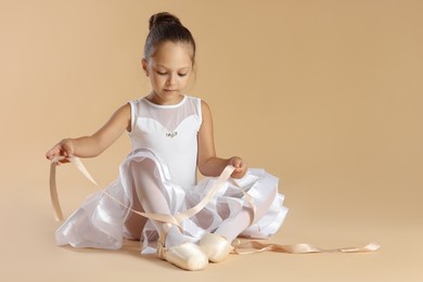 Little ballerina tying pointe shoes on beige background, space for text