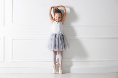 Little ballerina practicing dance moves near white wall indoors