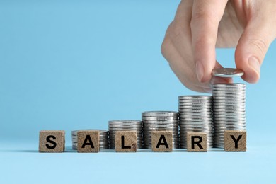 Photo of Woman putting coins and word Salary made of wooden cubes on light blue background, closeup. Space for text