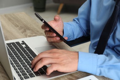 Photo of Businessman with smartphone using laptop at wooden table, closeup. Modern technology