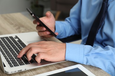 Photo of Businessman with smartphone using laptop at wooden table, closeup. Modern technology