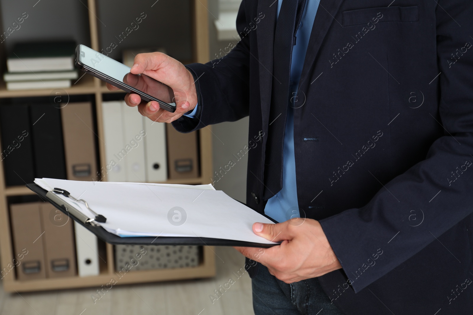 Photo of Businessman with clipboard using smartphone in office, closeup. Modern technology