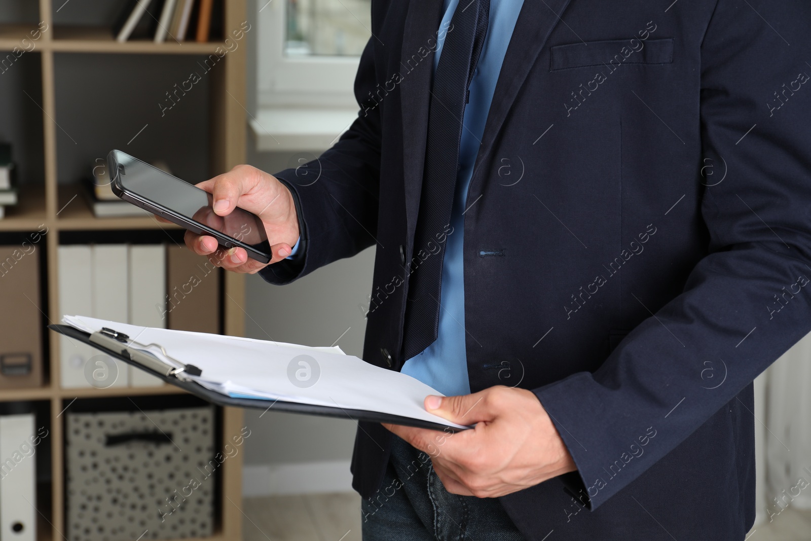 Photo of Businessman with clipboard using smartphone in office, closeup. Modern technology