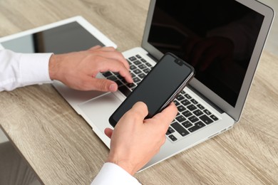 Photo of Businessman with smartphone using laptop at wooden table, closeup. Modern technology