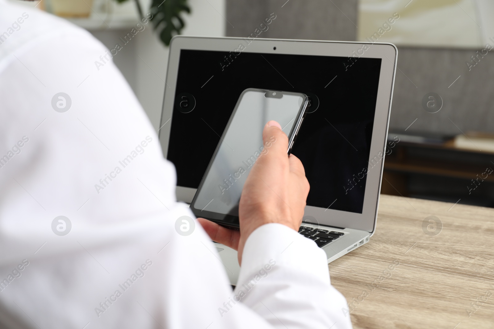 Photo of Businessman with smartphone using laptop at wooden table, closeup. Modern technology