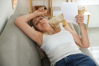 Photo of Menopause. Woman waving paper sheet to cool herself during hot flash on sofa at home