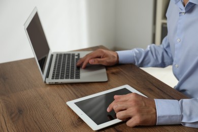 Photo of Businessman using laptop and tablet at wooden table, closeup. Modern technology