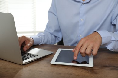 Photo of Businessman using laptop and tablet at wooden table, closeup. Modern technology