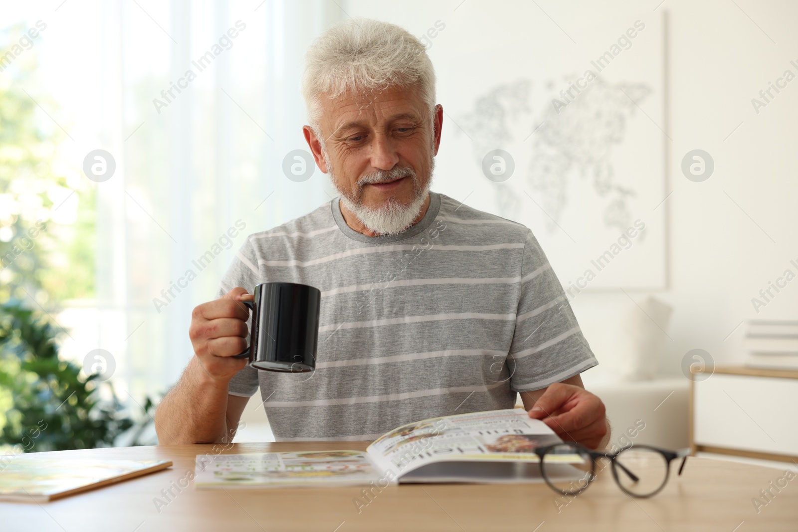 Photo of Senior man with cup of drink reading magazine at table indoors