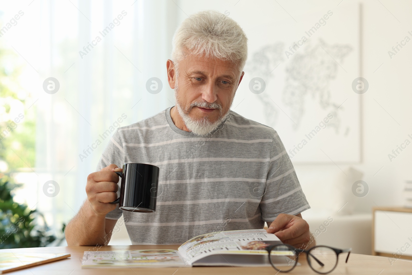 Photo of Senior man with cup of drink reading magazine at table indoors